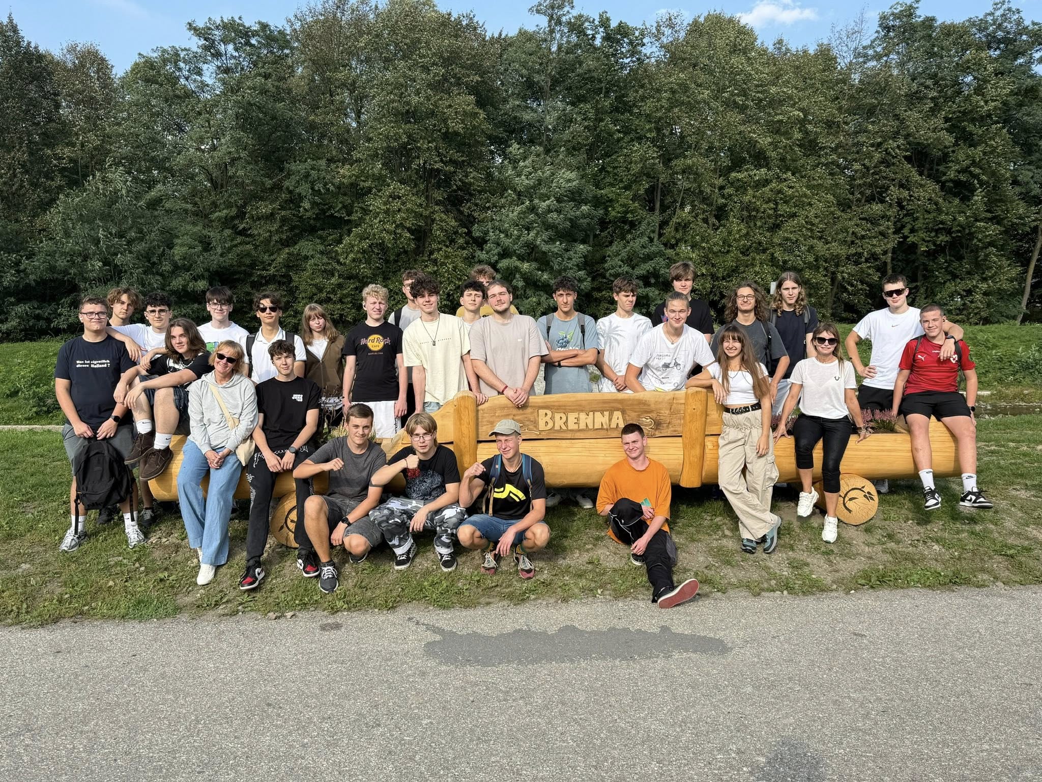 Część uczestników wycieczki z opiekunkami – zdjęcie grupowe, w tle las; na poziomo położonym drewnianym pniu/balu napis: Brenna