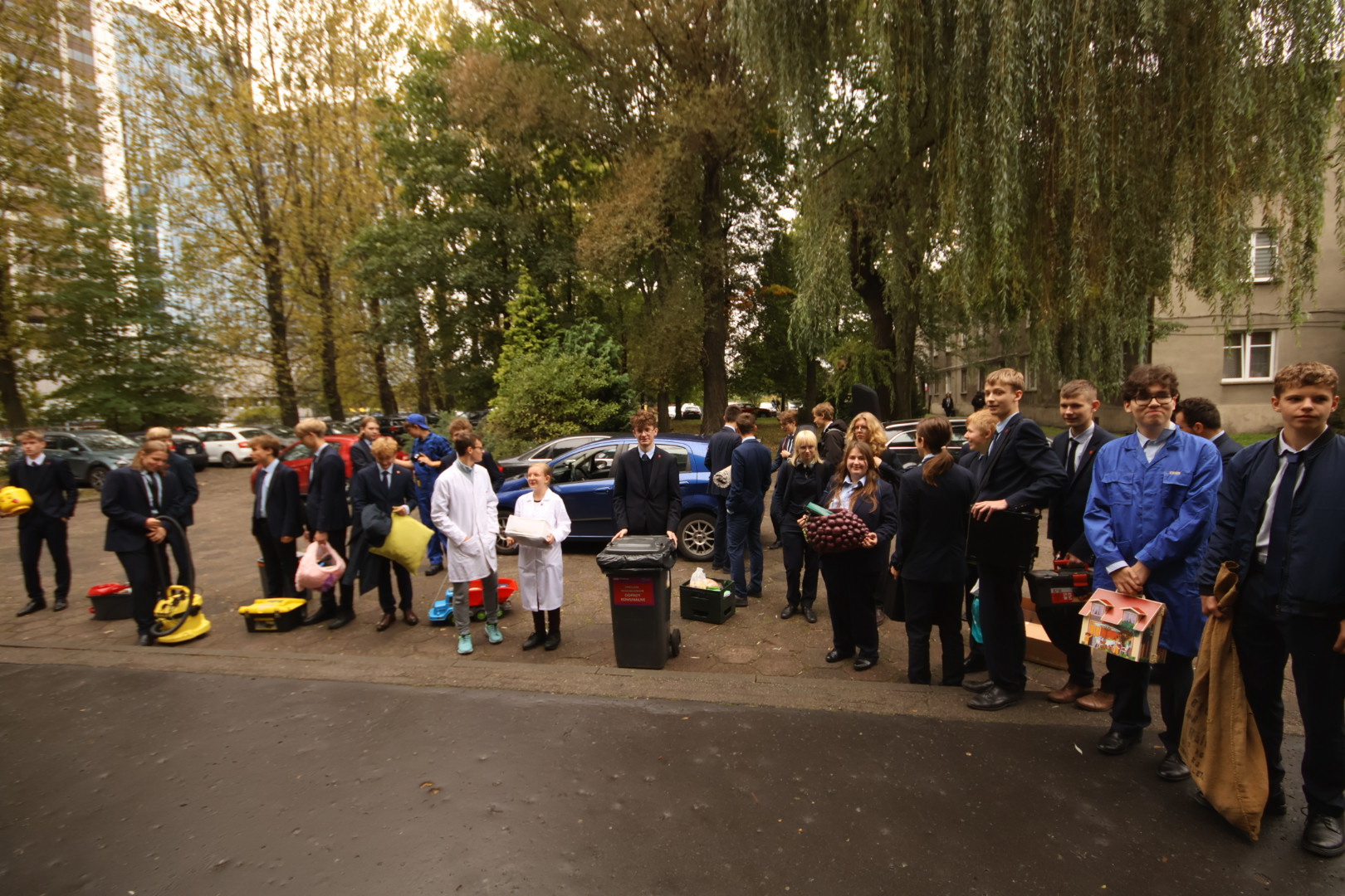 Uczennice i uczniowie przed szkołą w trakcie prezentacji różnych gadżetów zamiast tradycyjnych plecaków