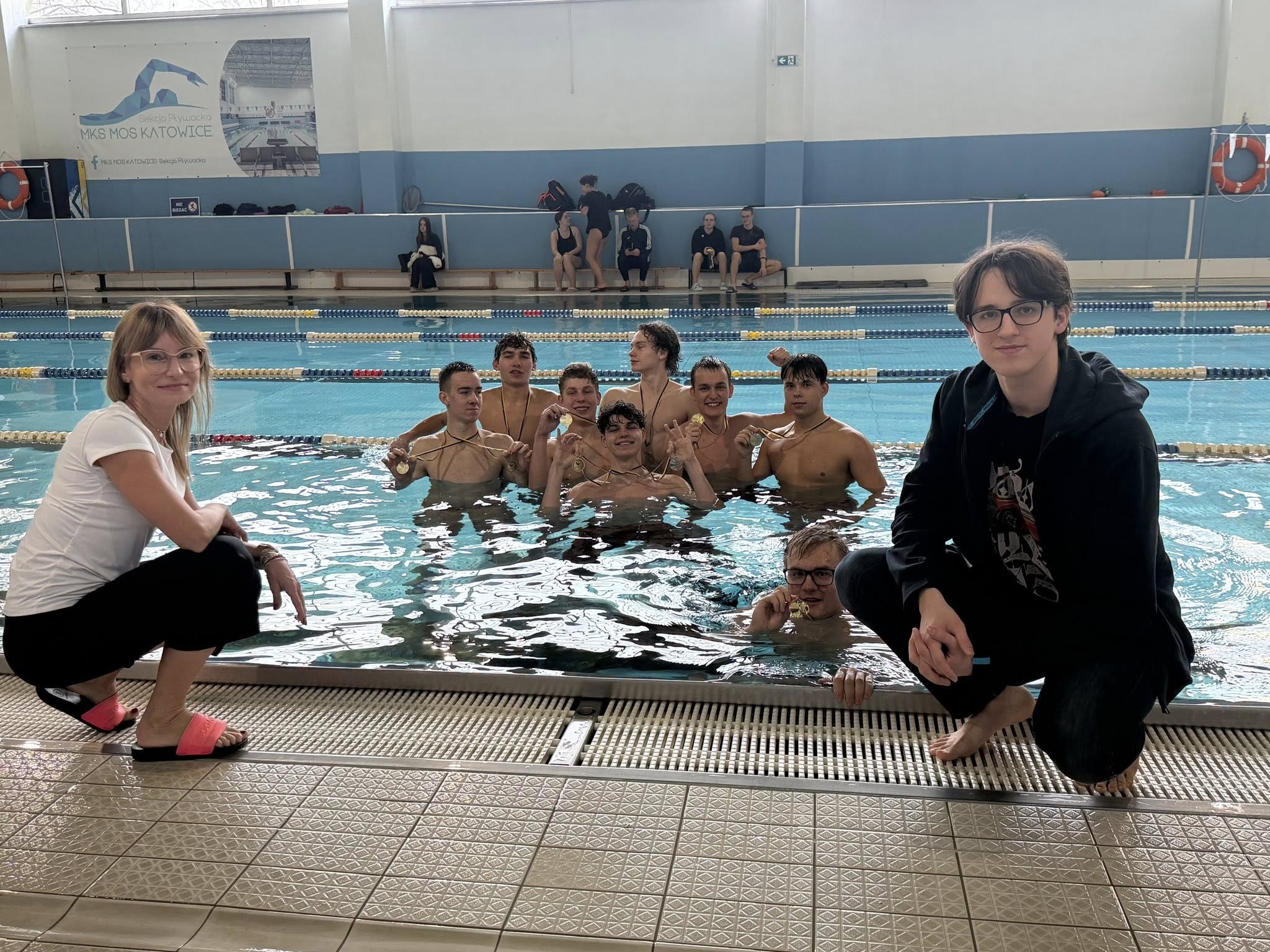 Sztafeta pływacka Śl.TZN, zdobywcy 1 miejsca z medalami, zanurzeni w wodzie basenu; na brzegu: z lewej opiekunka reprezentacji szkoły, z prawej zawodnik rezerwowy