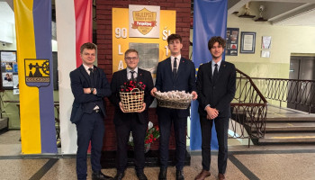 przedstawiciel SU przed tablicą pamiątkową w holu szkoły z koszem kwiatów i słodkości z okazji Dnia Kobiet