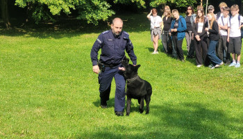 Funkcjonariusz Policji z psem szkolonym do łapania przestępców, z boku grupa zawodników obserwująca pokaz 