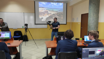 Uczniowie w sali komputerowej podczas zajęć, przy tablicy nauczyciel z kamerą