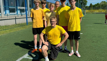 drużyna 1B – 7 zawodników w żółtych koszulkach sportowych; kapitan z przodu, w pozycji klęczącej na jedno kolano, reszta stoi; tło boisko