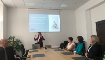 Warsztat dla opiekunów; widoczny na ekranie napis: Wypalenie zawodowe kryzys czy wyzwanie; prowadząca stoi i gestykuluje, uczestnicy siedzą wokół stołu