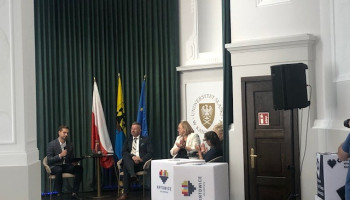 Rozmowa moderowana: moderator zadający pytanie i uczestnicy (widoczne 3 osoby); w tle flagi Polski, Katowic i Unii Europejskiej